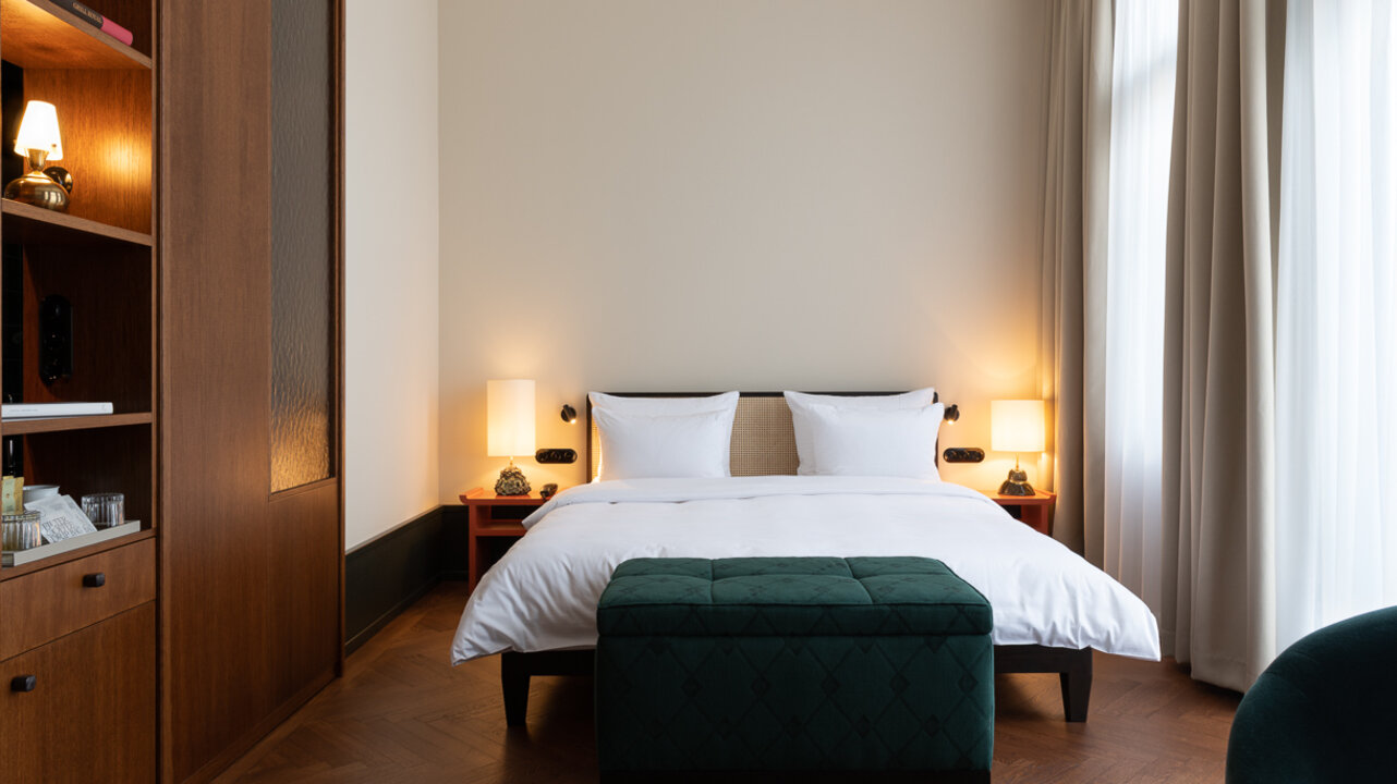 Der Schlafbereich in einem großen Hotelzimmer im Château Royal Berlin mit heller Fensterfront und einem großen Holzschrank.