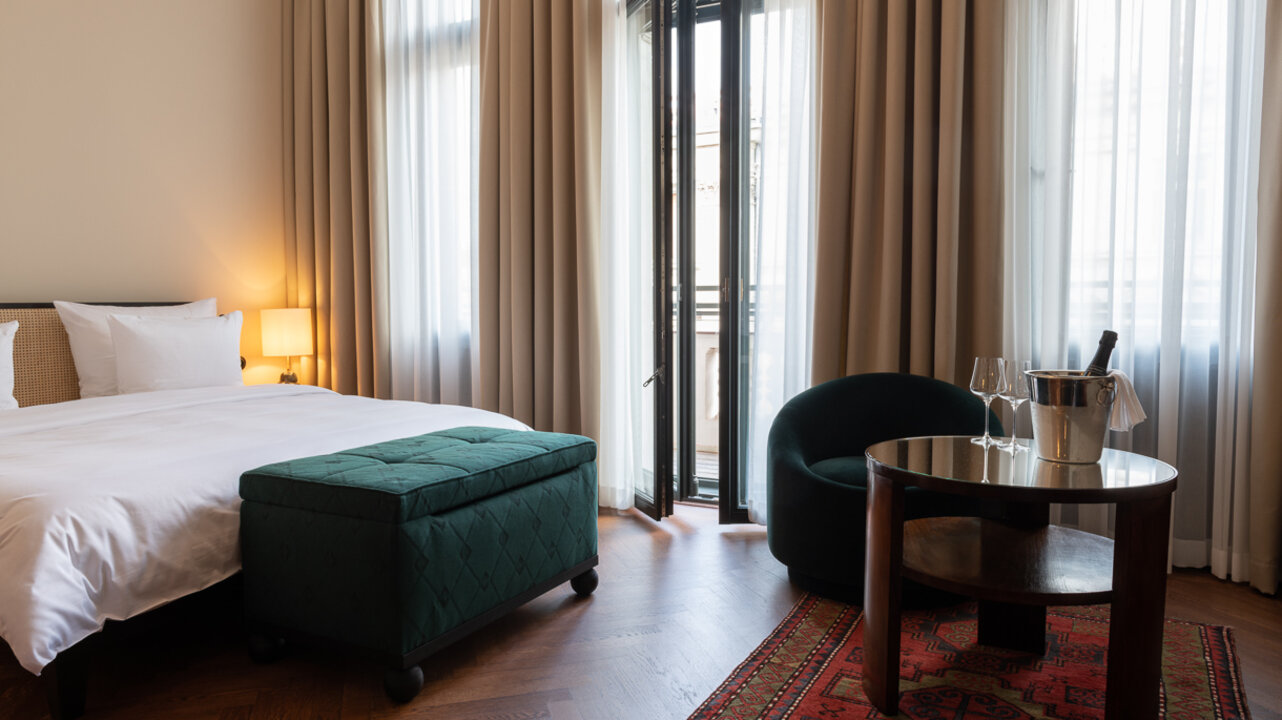 Blick auf das große Bett und die helle Fensterfront eines großen Hotelzimmers mit Balkon im Hotel Château Royal Berlin.