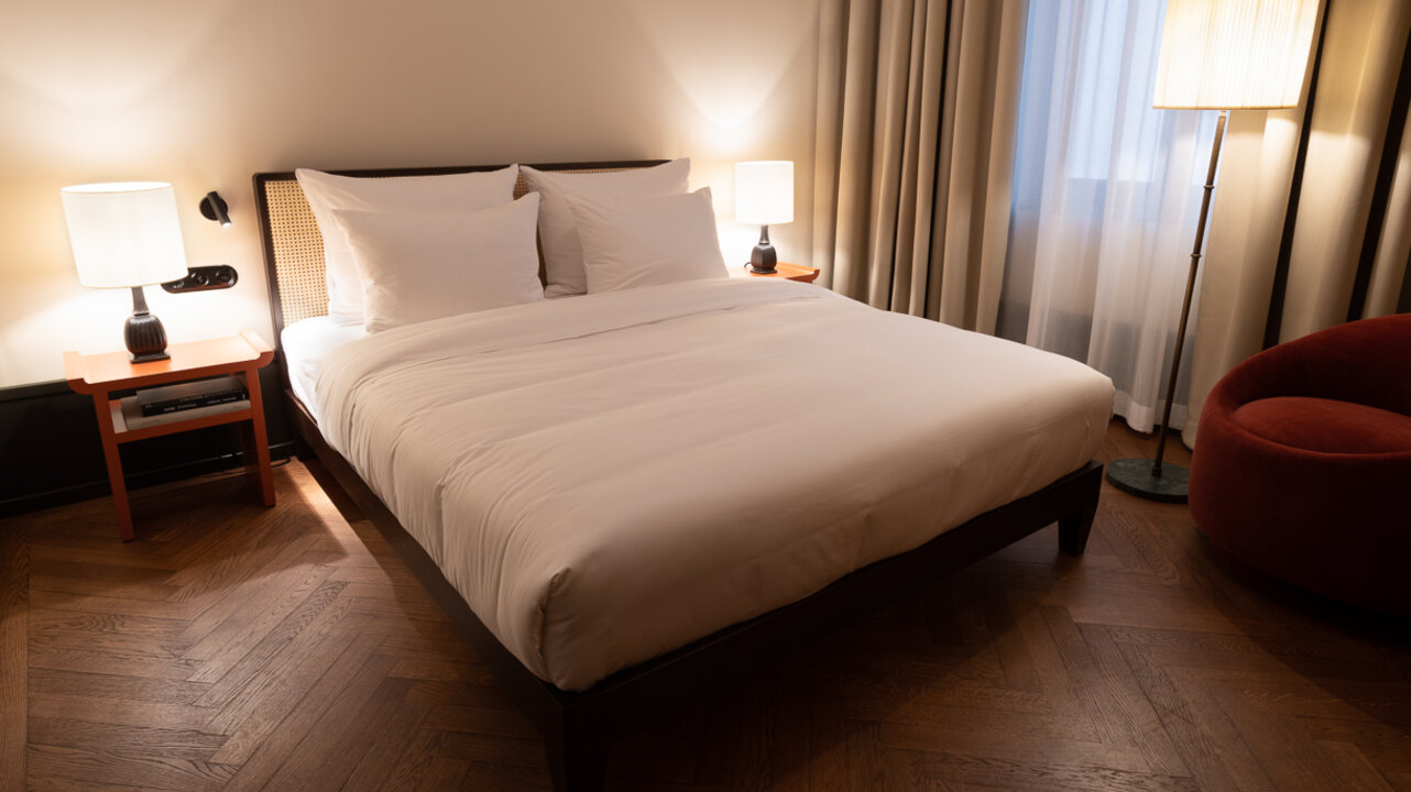 Blick auf ein Doppelbett in einem kleinen Hotelzimmer des Château Royal Berlin mit zwei Nachtkommoden und einem Fenster.