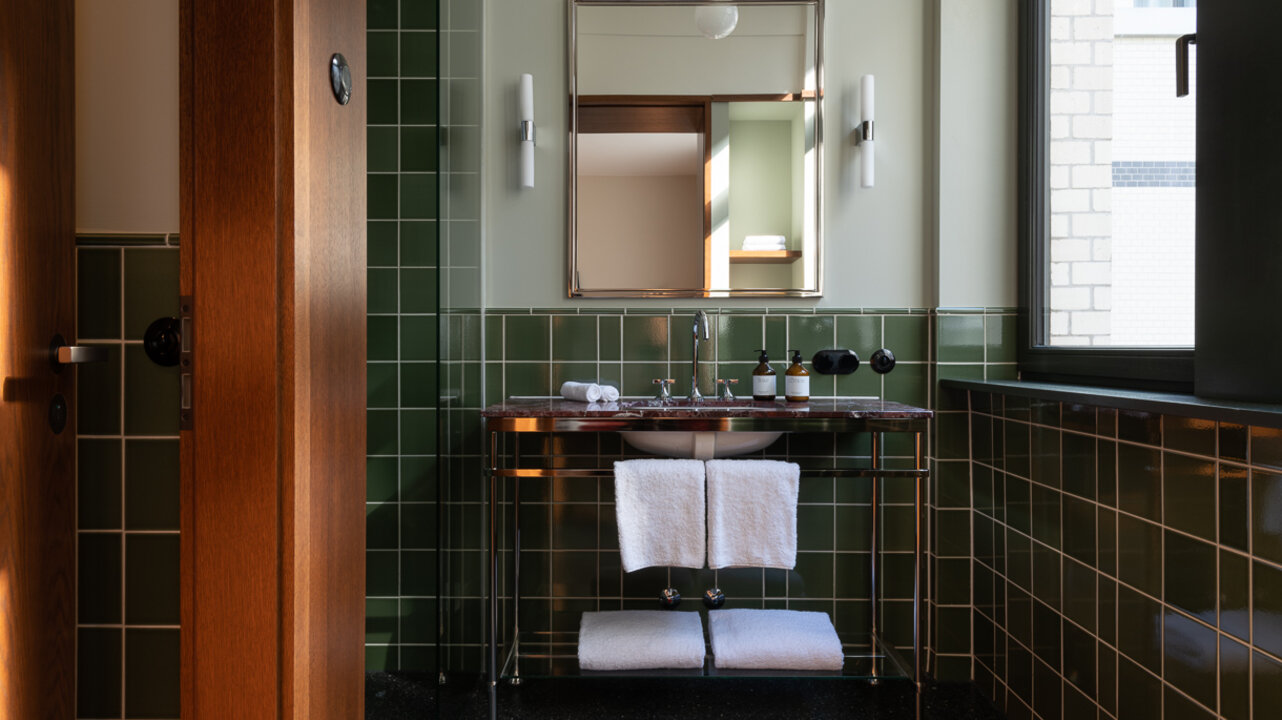 Blick auf das Waschbecken mit Spiegel neben dem Fenster in einem Bad des Châteu Royal Berlin, mit dunklen Fliesen.