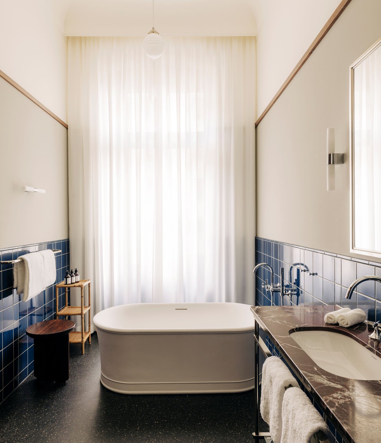 Blick in ein mit Tageslicht ausgestattets Bad mit dunkelblauen Fliesen, freistehender Badewanne und Marmorwaschbecken im Boutiquehotel Château Royal Berlin