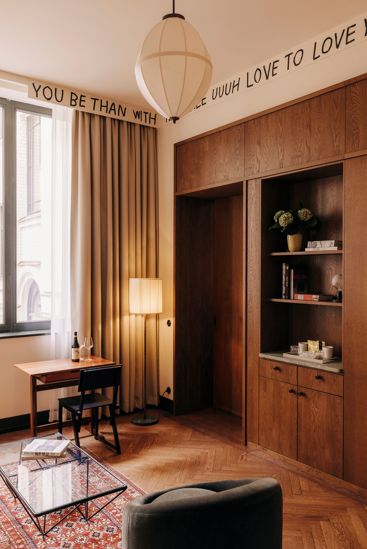 Wohnbereich eines Hotelzimmers im Boutiquehotel Château Royal Berlin ausgestattet mit einem großen Fenster, vor dem ein kleiner Schreibtisch steht, einer gemütlichen Sitzgelegenheit und einer großen Schrankwand in dunklen Holztönen