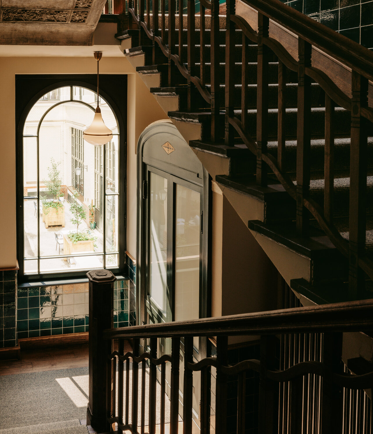 Blick auf das große halbrunde Fenster im historischen Treppenhaus des Boutiquehotel Château Royal Berlin mit dunkler Holztreppe und dunkelgrünen Wandfliesen